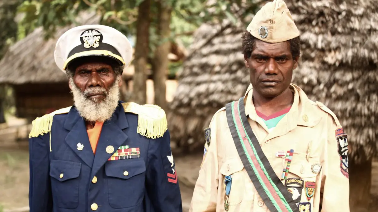Dos hombres melanesios de pie frente a chozas tradicionales, vestidos con uniformes militares simbólicos decorados con insignias y medallas, característicos de los cultos cargo de Melanesia